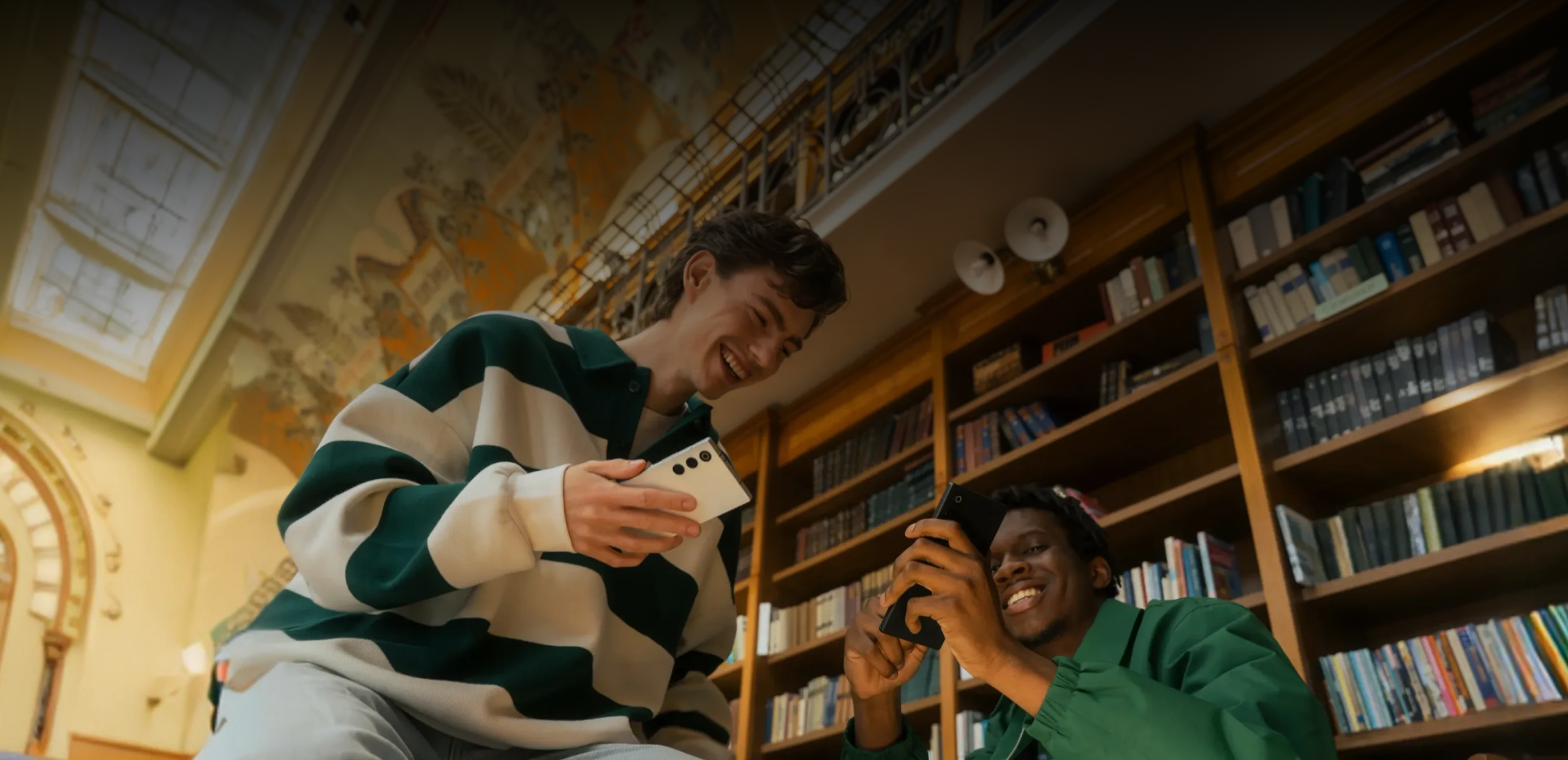 Two young men smiling and looking at their smartphones in a library with bookshelves and a decorated ceiling.